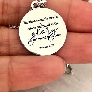 May include: A circular silver-toned metal pendant necklace featuring the engraved black text: Yet what we suffer now is nothing compared to the glory he will reveal to us later Romans 8:18. The pendant hangs from a delicate silver-toned chain.