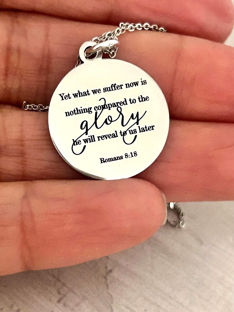 May include: A circular silver-toned metal pendant necklace featuring the engraved black text: Yet what we suffer now is nothing compared to the glory he will reveal to us later Romans 8:18. The pendant hangs from a delicate silver-toned chain.