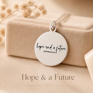 May include: A silver-colored pendant necklace with a round charm. The charm has the words "hope and a future" and "Jeremiah 29:11" in black script. The necklace is displayed on a beige jewelry box. The words "Hope & a Future" are at the bottom.