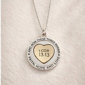 May include: Silver-tone necklace with a circular pendant. The pendant features a gold heart in the center with the text "1 COR 13:13". The outer ring has the words "FAITH, HOPE AND LOVE" and "AND NOW THESE THREE REMAIN".