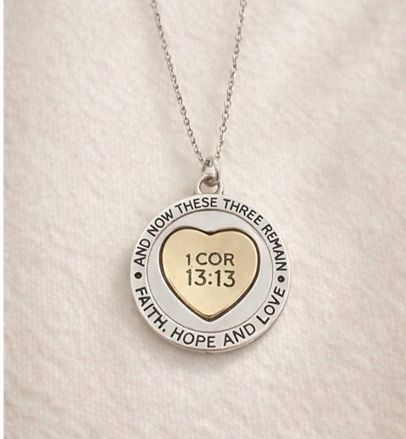 May include: Silver-tone necklace with a circular pendant. The pendant features a gold heart in the center with the text "1 COR 13:13". The outer ring has the words "FAITH, HOPE AND LOVE" and "AND NOW THESE THREE REMAIN".