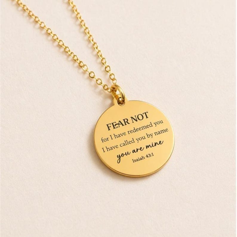 May include: A gold-toned circular pendant necklace featuring a delicate chain and a round charm engraved with the black text: FEAR NOT for I have redeemed you I have called you by name you are mine Isaiah 43:1. The piece has a polished finish.