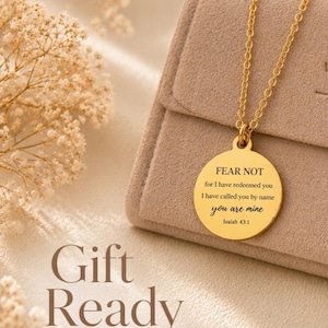 May include: A gold-toned circular pendant necklace featuring the engraved text FEAR NOT for I have redeemed you I have called you by name you are mine Isaiah 43:1. The pendant hangs from a delicate gold-toned chain resting on a beige fabric surface.