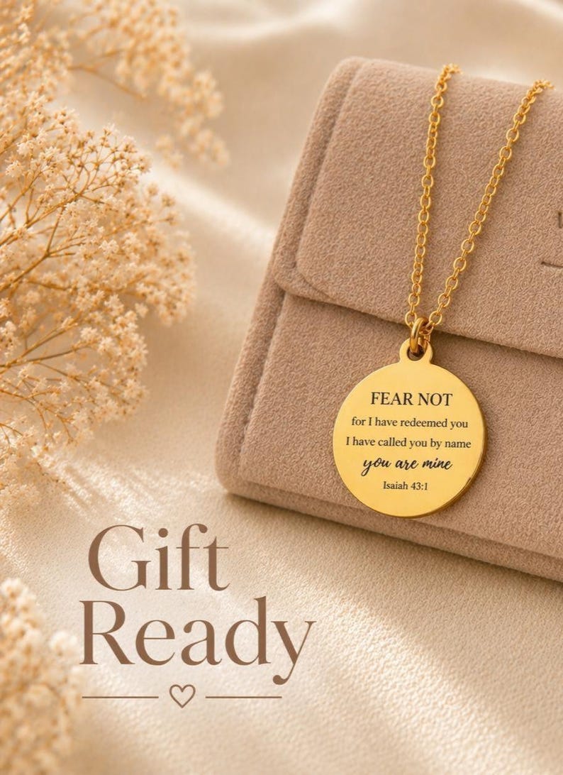 May include: A gold-toned circular pendant necklace featuring the engraved text FEAR NOT for I have redeemed you I have called you by name you are mine Isaiah 43:1. The pendant hangs from a delicate gold-toned chain resting on a beige fabric surface.