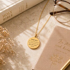 May include: A gold-toned circular pendant necklace featuring the engraved text FEAR NOT for I have redeemed you I have called you by name you are mine Isaiah 43:1. The necklace is displayed alongside a Holy Bible, eyeglasses, and a notebook.