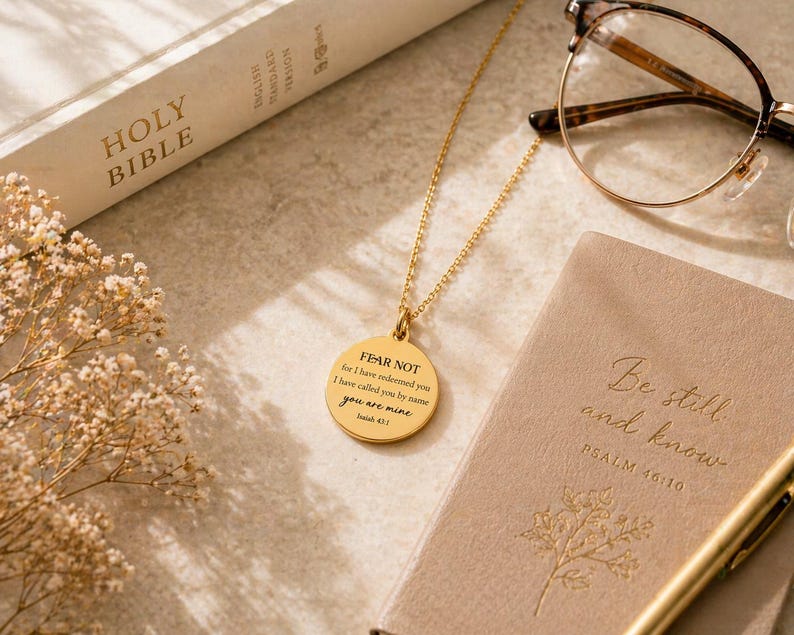 May include: A gold-toned circular pendant necklace featuring the engraved text FEAR NOT for I have redeemed you I have called you by name you are mine Isaiah 43:1. The necklace is displayed alongside a Holy Bible, eyeglasses, and a notebook.