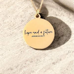 May include: A gold-colored pendant necklace with a round charm. The charm has the words "hope and a future" and "Jeremiah 29:11" in black script. The necklace chain is also gold-colored.