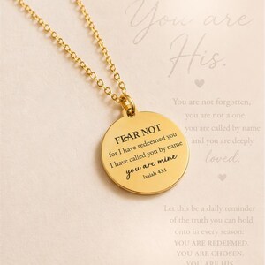 May include: A gold-toned circular pendant necklace featuring the engraved text FEAR NOT for I have redeemed you I have called you by name you are mine Isaiah 43:1. The pendant hangs from a delicate gold-toned chain against a light background.