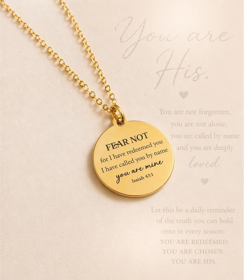 May include: A gold-toned circular pendant necklace featuring the engraved text FEAR NOT for I have redeemed you I have called you by name you are mine Isaiah 43:1. The pendant hangs from a delicate gold-toned chain against a light background.