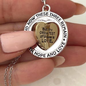 May include: Silver-toned necklace with a circular pendant. The pendant features a gold-colored heart inside a silver ring. The ring is inscribed with the words "NOW THESE THREE REMAIN" and "WITH HOPE AND LOVE." The heart has the inscription "But the GREATEST of these is LOVE."