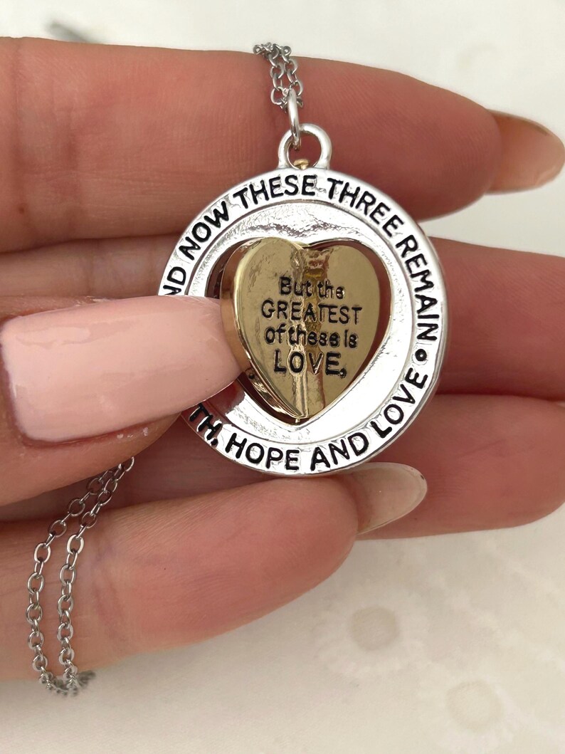 May include: Silver-toned necklace with a circular pendant. The pendant features a gold-colored heart inside a silver ring. The ring is inscribed with the words "NOW THESE THREE REMAIN" and "WITH HOPE AND LOVE." The heart has the inscription "But the GREATEST of these is LOVE."