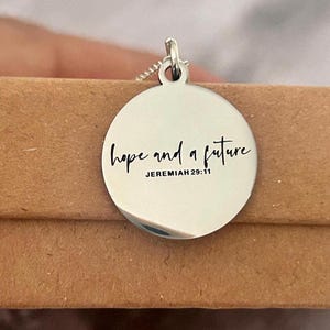 May include: Silver-colored pendant necklace with a round charm. The charm is engraved with the words "hope and a future" and the reference "JEREMIAH 29:11" in a black script font. The necklace is displayed on a brown surface.
