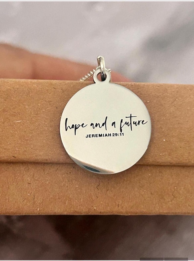 May include: Silver-colored pendant necklace with a round charm. The charm is engraved with the words "hope and a future" and the reference "JEREMIAH 29:11" in a black script font. The necklace is displayed on a brown surface.