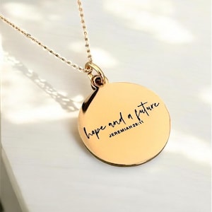 Puede incluir: Collar dorado con un colgante circular. El colgante presenta la inscripción "hope and a future" y la referencia "JEREMIAH 29:11". La cadena es delicada y el colgante es reflectante.