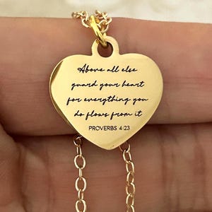 Könnte beinhalten: Eine goldfarbene herzförmige Anhänger-Halskette. Der Anhänger trägt die Inschrift "Above all else guard your heart for everything you do flows from it PROVERBS 4:23" in schwarzer Schrift. Die Halskette ist ebenfalls goldfarben.