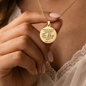 Op de afbeelding: Een goudkleurige ronde hanger aan een ketting met de gegraveerde tekst: WHEN MY HEART IS OVERWHELMED; LEAD ME TO The Rock THAT IS HIGHER THAN I Psalm 61:2. De hanger hangt aan een fijne goudkleurige ketting met een gepolijste metalen afwerking.