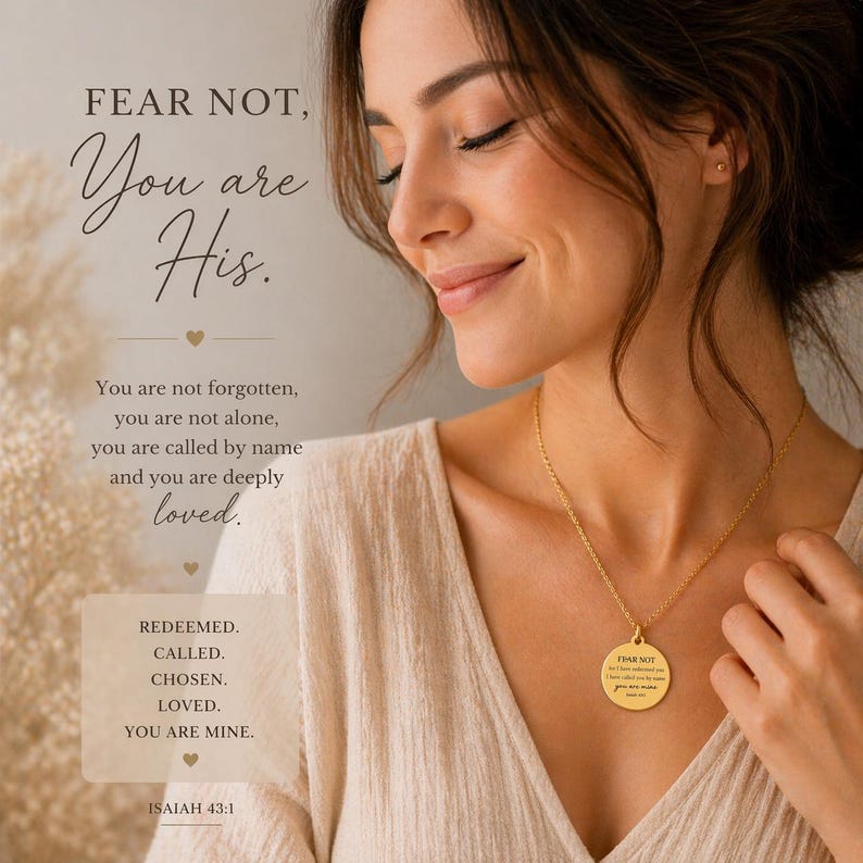 May include: A gold-toned circular pendant necklace featuring engraved text that reads FEAR NOT, but I have redeemed you, I have called you by name, you are mine, Isaiah 43:1. The pendant hangs from a delicate gold chain against a light-colored fabric.