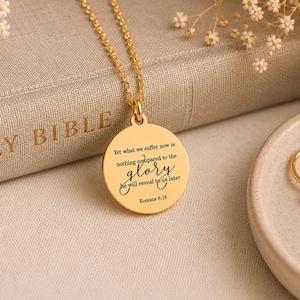 May include: A gold-toned circular pendant necklace featuring a delicate chain and an engraved inscription that reads: Yet what we suffer now is nothing compared to the glory he will reveal to us later Romans 8:18 in a mix of serif and script fonts.