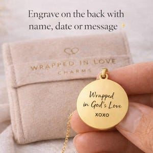 May include: A gold-colored pendant necklace with the inscription "Wrapped in God's Love XOXO" on a round charm. The necklace is displayed with a beige pouch that has the words "Wrapped in Love Charms" printed on it. The text "Engrave on the back with name, date or message" is also visible.