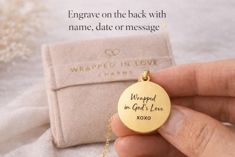 May include: A gold-colored pendant necklace with the inscription "Wrapped in God's Love XOXO" on a round charm. The necklace is displayed with a beige pouch that has the words "Wrapped in Love Charms" printed on it. The text "Engrave on the back with name, date or message" is also visible.