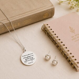 May include: A silver necklace with a round pendant engraved with the text 'Yet what we suffer now is nothing compared to the glory he will reveal to us later Romans 8:18' and a pair of silver stud earrings featuring small white pearls.