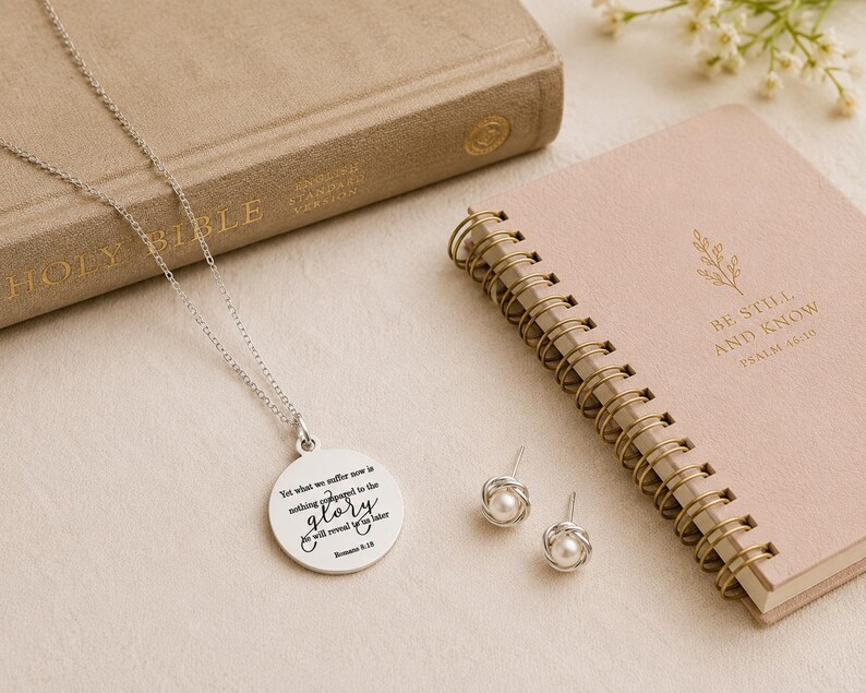 May include: A silver necklace with a round pendant engraved with the text 'Yet what we suffer now is nothing compared to the glory he will reveal to us later Romans 8:18' and a pair of silver stud earrings featuring small white pearls.