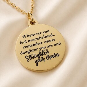 May include: A gold-colored pendant necklace with a round charm. The charm has the engraved text: "Whenever you feel overwhelmed... remember whose daughter you are and Straighten your crown."