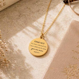 May include: A gold-toned circular pendant necklace featuring the engraved text FEAR NOT for I have redeemed you I have called you by name you are mine Isaiah 43:1. The pendant hangs from a delicate gold-toned chain with a polished finish.