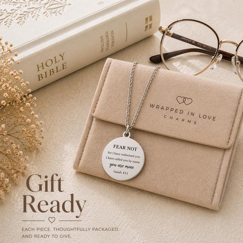 May include: A silver-toned circular pendant necklace featuring the engraved text FEAR NOT for I have redeemed you I have called you by name you are mine Isaiah 43:1. The necklace rests on a beige velvet pouch with the logo WRAPPED IN LOVE CHARMS.