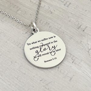 May include: A silver-toned circular pendant necklace featuring the engraved text: Yet what we suffer now is nothing compared to the glory he will reveal to us later Romans 8:18. The pendant hangs from a delicate matching silver-toned metal chain.