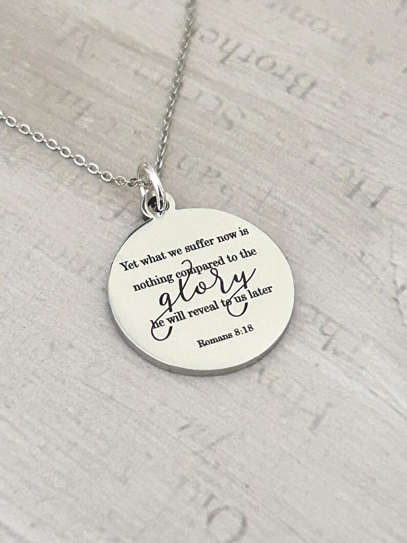 May include: A silver-toned circular pendant necklace featuring the engraved text: Yet what we suffer now is nothing compared to the glory he will reveal to us later Romans 8:18. The pendant hangs from a delicate matching silver-toned metal chain.