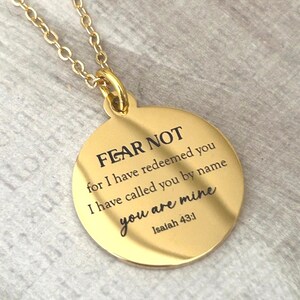 May include: A gold-toned circular pendant necklace featuring the engraved text FEAR NOT for I have redeemed you I have called you by name you are mine Isaiah 43:1. The pendant hangs from a matching gold-toned chain with a polished metallic finish.