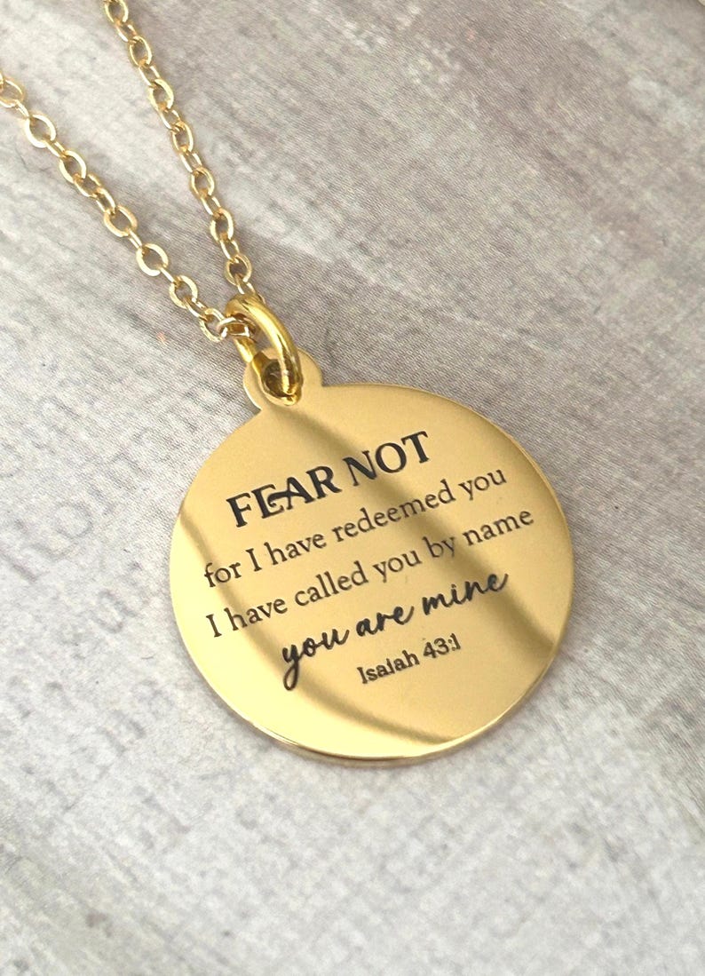 May include: A gold-toned circular pendant necklace featuring the engraved text FEAR NOT for I have redeemed you I have called you by name you are mine Isaiah 43:1. The pendant hangs from a matching gold-toned chain with a polished metallic finish.