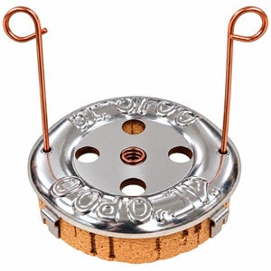 Op de afbeelding: Een cirkelvormig metalen apparaat met een glanzende zilveren afwerking en in reliëf aangebrachte tekst "GLO-CO" en "AIR-O-POO". Het heeft vier ronde uitsparingen, een centrale veer en twee rechtopstaande koperkleurige draadlussen op een kurken voet.