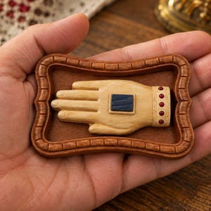 May include: A small rectangular wooden frame containing a carved light-colored wooden hand. The hand features a dark blue square inlay on the palm and a cuff decorated with five small red circular accents. The frame has a brown textured background.