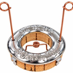 Könnte beinhalten: Ein runder Korksockel mit einem silberfarbenen Metallring, der den geprägten Text "D.V. OPOO" trägt. Zwei vertikale Kupferdrahtschlaufen ragen aus dem Ring nach oben, und ein kleiner Kupferdraht-Federmechanismus befindet sich in der mittleren Öffnung.