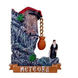 Könnte beinhalten: Souvenir mit einem Kloster auf einer Felsklippe, mit rotem Dach und einer kleinen Mönchsfigur. Auf dem Sockel steht "METEORA" in goldfarbenen Buchstaben. Eine goldene Kette hängt vom Gebäude zu einem braunen Beutel.