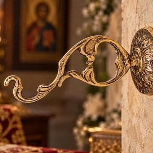 Puede incluir: Un gancho de pared de latón con un diseño curvo y ornamentado con una placa base circular. El metal tiene un acabado pulido y reflectante con detalles en relieve intrincados a lo largo del brazo y la placa de montaje, fijado con un tornillo.
