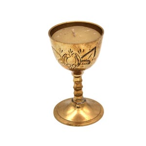 Puede incluir: Un portavelas de color dorado con un diseño floral grabado en la copa. El portavelas tiene una base redonda, un tallo con un diseño acanalado y una copa que contiene una vela beige. El portavelas es de metal.