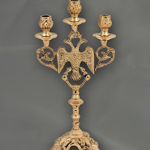 Puede incluir: Un candelabro dorado con tres candeleros. El diseño central presenta un emblema de águila bicéfala. La base es ornamentada y el estilo general es decorativo y detallado.