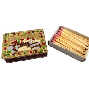 Puede incluir: Una caja de cerillas decorativa con tapa dorada que presenta un diseño de paisaje detallado con detalles en rojo, verde y blanco. Una caja de cerillas con puntas rosas está al lado.
