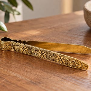 May include: A gold-toned metal tong featuring an intricate embossed floral pattern along the exterior of the handle. The tool has a spring-loaded hinge at the top and two long, flat, pointed arms that taper toward the ends for gripping small items.