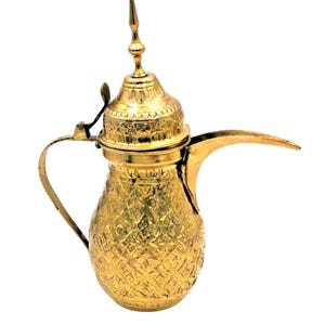 Puede incluir: Una cafetera de metal dorada con un diseño ornamentado y una tapa. La cafetera tiene un pico curvo y un asa.