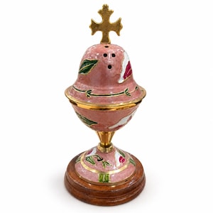 May include: A decorative metal censer featuring a pink enameled body with hand-painted green leaf and white floral motifs. The piece includes a gold-toned cross finial at the top, a brass-colored stem, and a circular wooden base for stability.