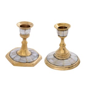 May include: Two brass candle holders with a mother-of-pearl inlay. One has a hexagonal base, the other a round base. The candle holders have a gold finish and are designed to hold a single candle. They are set against a white background.