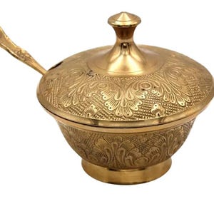 May include: A gold-coloured, lidded serving bowl with an ornate floral pattern. The bowl includes a matching spoon resting on the rim. The lid has a small, rounded handle. The bowl sits on a small, round base.