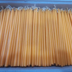 Puede incluir: Una caja llena de numerosas velas cónicas de color amarillo anaranjado. Cada vela tiene una mecha blanca en la parte superior. Las velas están dispuestas muy juntas, creando una apariencia densa y texturizada. La impresión general es de una gran cantidad de velas.