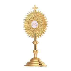 Puede incluir: Una custodia dorada con un diseño de rayos de sol. El elemento central presenta un círculo blanco con las letras "JHS". La custodia tiene una cruz en la parte superior y una base decorativa.