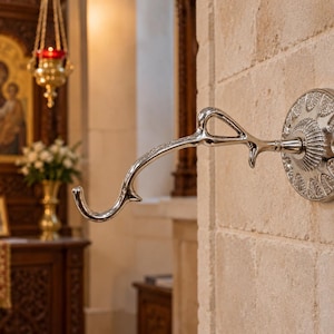 May include: A polished silver-toned metal wall hook featuring a curved, branch-like arm extending from a circular base plate. The base plate is decorated with a repeating embossed leaf pattern and is mounted on a light-colored stone wall.
