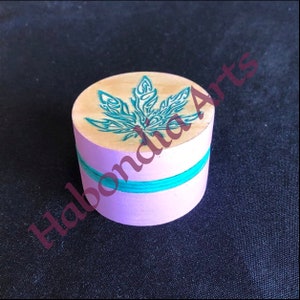 Puede incluir: Una pequeña caja de madera con una tapa rosa claro y un diseño de hoja de marihuana verde en la parte superior. La caja tiene una banda de color turquesa alrededor del medio.